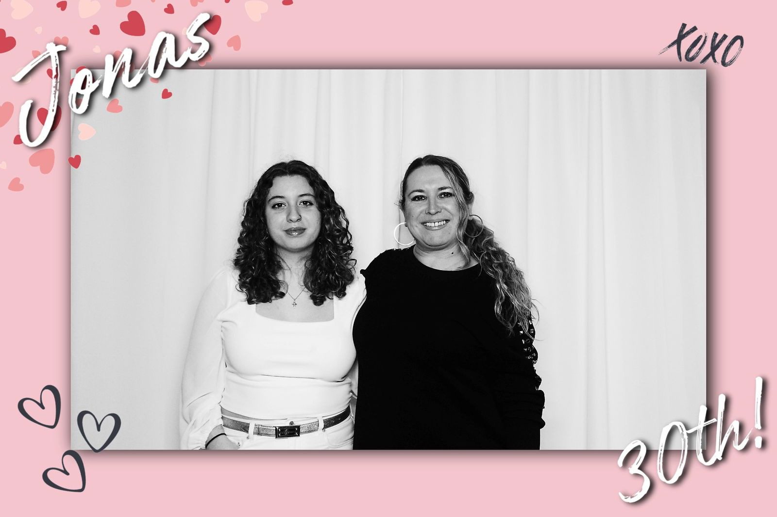 Two women standing side by side against a light backdrop, one in white and one in black.