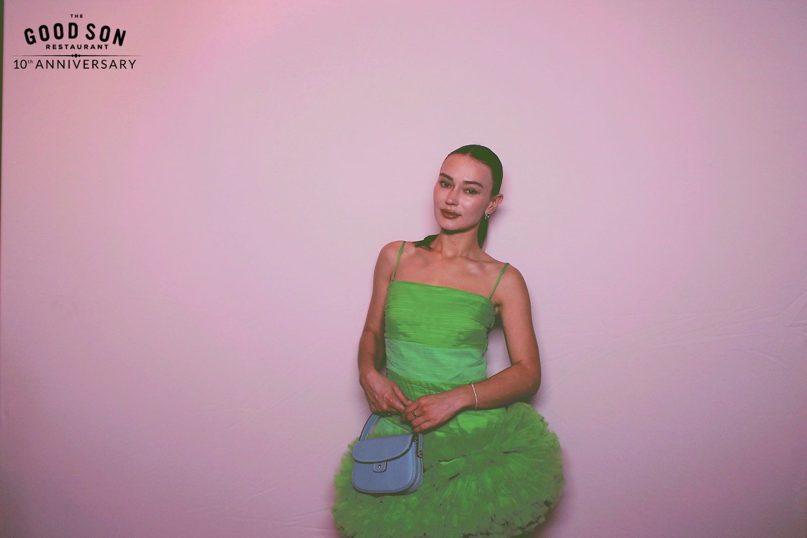 Woman in a vibrant green dress posing with a blue handbag against a pink background.